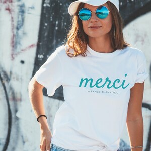 May include: White t-shirt with the word "merci" in teal blue script and the text "a fancy thank you" in black below. The model is wearing the shirt with blue jeans and a white cap.