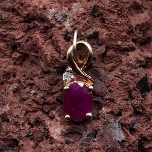 May include: A gold pendant featuring a central oval-cut deep pink ruby gemstone held by four prongs, accented by a small clear round-cut diamond set above the ruby, with a polished gold bail at the top for attaching to a chain or necklace.