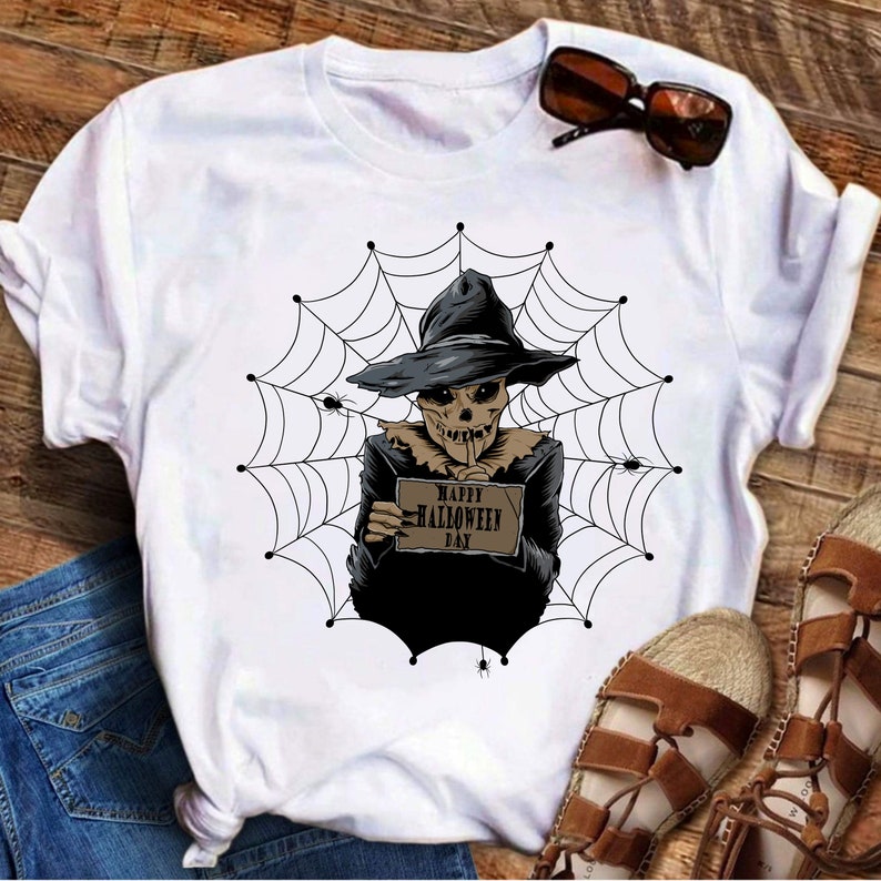 K&ouml;nnte beinhalten: Wei&szlig;es T-Shirt mit einem Aufdruck eines Skeletts, das einen Hexenhut tr&auml;gt und ein Schild mit der Aufschrift "Happy Halloween Day" in einem Spinnennetz h&auml;lt.