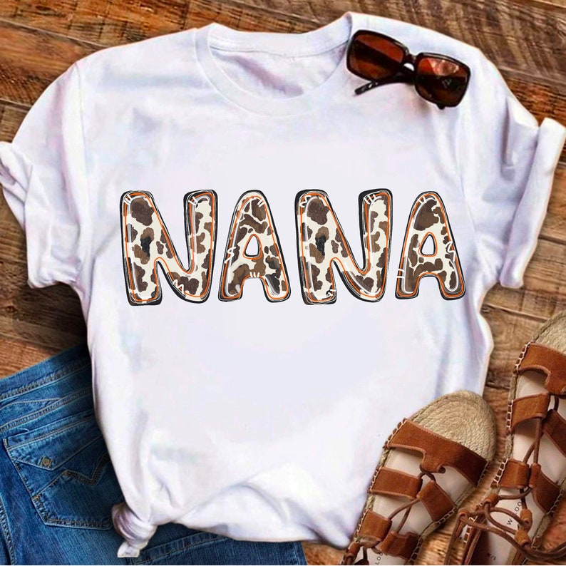 Puede incluir: Camiseta blanca con la palabra "NANA" en letras de estampado de leopardo marr&oacute;n y blanco.