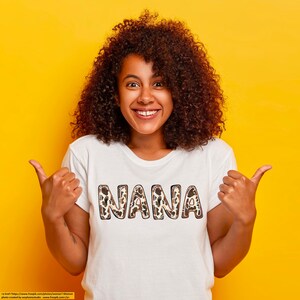 Puede incluir: Una mujer con una camiseta blanca con la palabra "NANA" impresa en letras de estampado de leopardo marr&oacute;n y negro. Est&aacute; sonriendo y haciendo un pulgar hacia arriba con ambas manos.