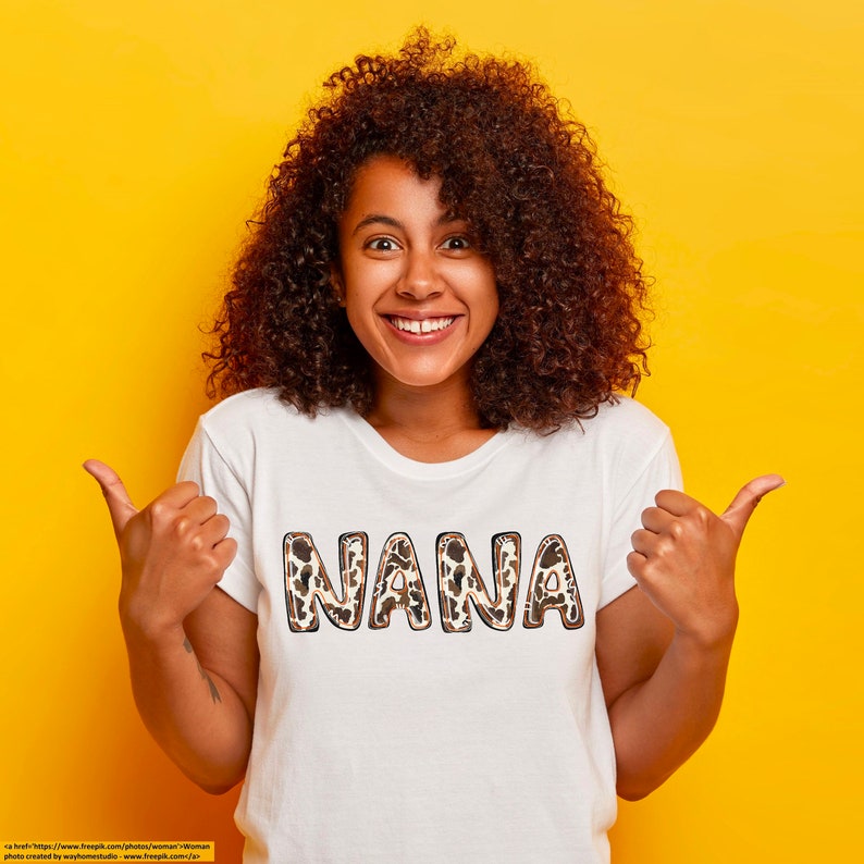 Puede incluir: Una mujer con una camiseta blanca con la palabra "NANA" impresa en letras de estampado de leopardo marr&oacute;n y negro. Est&aacute; sonriendo y haciendo un pulgar hacia arriba con ambas manos.