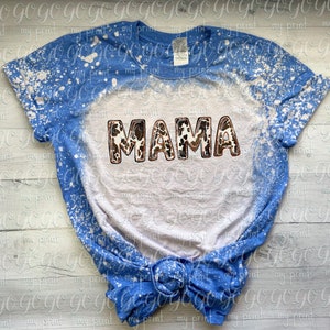 Puede incluir: Una camiseta azul y blanca blanqueada con la palabra "MAMA" en letras marrones y blancas de estampado de vaca.