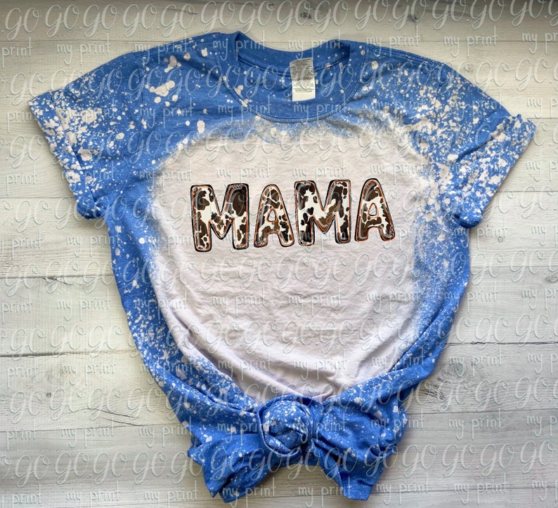 Puede incluir: Una camiseta azul y blanca blanqueada con la palabra "MAMA" en letras marrones y blancas de estampado de vaca.