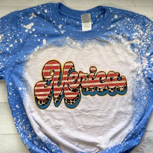 May include: A white t-shirt with a blue splatter design on the sleeves. The shirt has a red, white, and blue "Merica" graphic with a leopard print design.