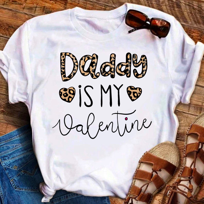 May include: White t-shirt with the text "Daddy is my Valentine" in black script. The word "Daddy" is in leopard print. There are two leopard print hearts around the word "my".