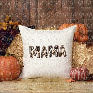 Puede incluir: Una almohada blanca con la palabra "MAMA" en letras de estampado animal marr&oacute;n, negro y blanco. La almohada est&aacute; sentada en un lecho de heno con calabazas y hojas de oto&ntilde;o a su alrededor.