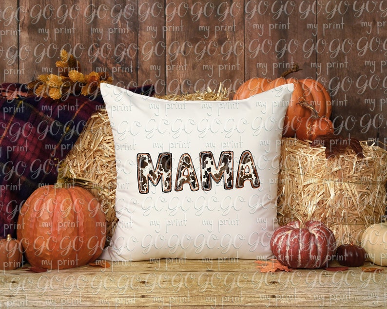 Puede incluir: Una almohada blanca con la palabra "MAMA" en letras de estampado animal marr&oacute;n, negro y blanco. La almohada est&aacute; sentada en un lecho de heno con calabazas y hojas de oto&ntilde;o a su alrededor.