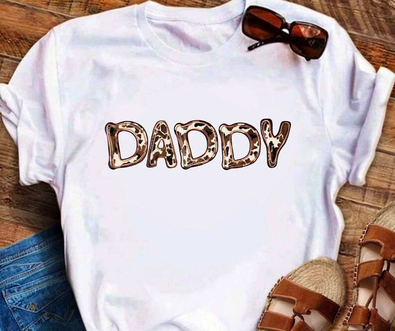 May include: White t-shirt with the word "DADDY" printed in brown and tan leopard print.