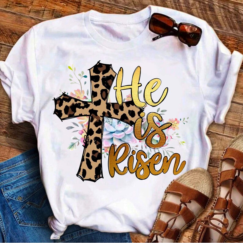 May include: White t-shirt with a leopard print cross and the text "He is risen" in gold lettering. The cross is surrounded by floral designs.