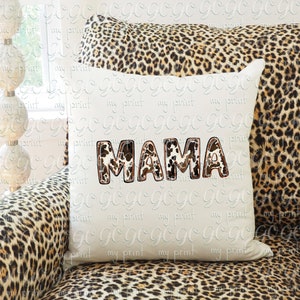Puede incluir: Una almohada blanca con la palabra "MAMA" escrita en letras de estampado de leopardo marr&oacute;n y blanco. La almohada est&aacute; sentada en un sof&aacute; de estampado de leopardo marr&oacute;n y negro.