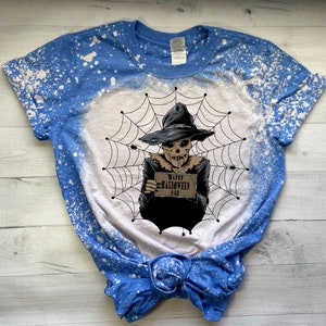 K&ouml;nnte beinhalten: Ein blaues und wei&szlig;es Batik-T-Shirt mit einem Design aus Bleichmittelspritzern. Das Shirt zeigt eine Grafik eines Skeletts, das einen Hexenhut tr&auml;gt und ein Schild mit der Aufschrift "Happy Halloween Day" h&auml;lt.