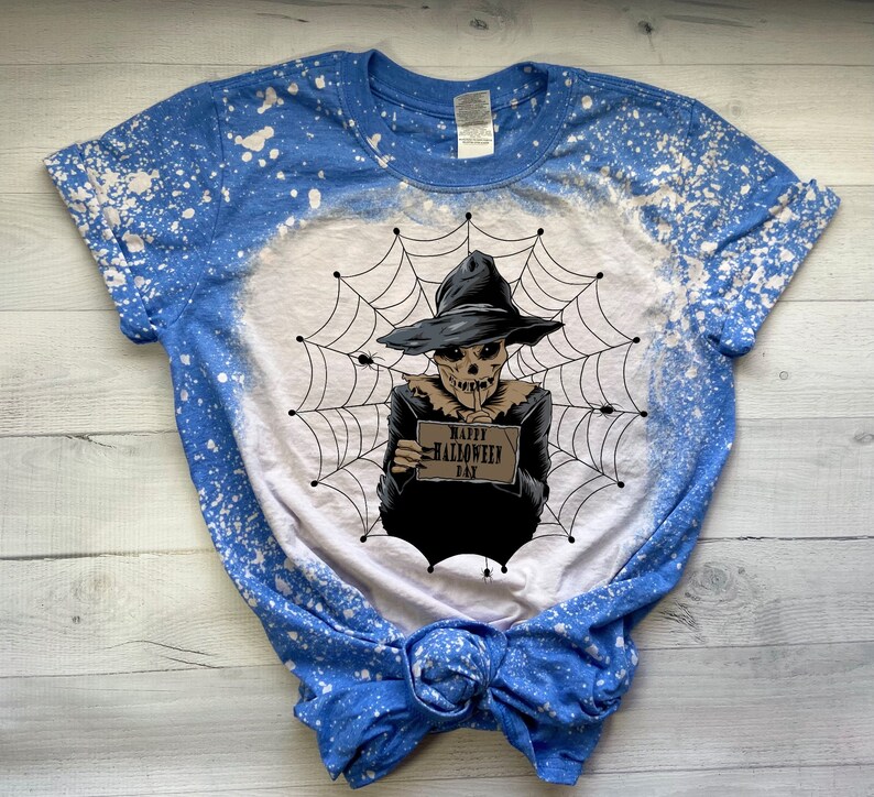 K&ouml;nnte beinhalten: Ein blaues und wei&szlig;es Batik-T-Shirt mit einem Design aus Bleichmittelspritzern. Das Shirt zeigt eine Grafik eines Skeletts, das einen Hexenhut tr&auml;gt und ein Schild mit der Aufschrift "Happy Halloween Day" h&auml;lt.
