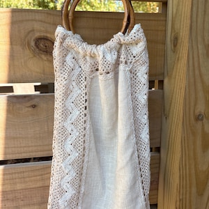 May include: A beige linen drawstring bag featuring intricate cream-colored crochet side panels with a zigzag pattern and two circular bamboo handles. The bag has a gathered top opening and a rectangular body made of plain woven fabric.