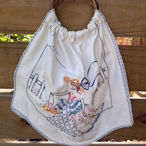 May include: A white fabric bag with a round bamboo handle featuring an embroidered design of a person wearing a wide-brimmed hat and a dress with blue and pink floral patterns. The bag has a blue scalloped edge trim and a gathered top opening.