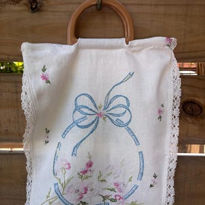 May include: A white fabric pouch with a rounded brown plastic handle, featuring an embroidered blue ribbon bow and pink floral bouquet design. The edges are finished with white lace trim, and the pouch is suspended from a metal hook.