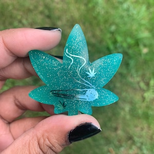 May include: A teal green glitter resin leaf with a white outline of a marijuana leaf and a joint with smoke coming out of it.