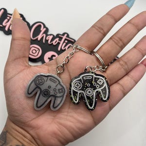 May include: Two controller-shaped keychains. One is light gray with black detailing, the other is black with green and white accents. Both have silver key rings and chains. The keychains are held up in front of a white background.