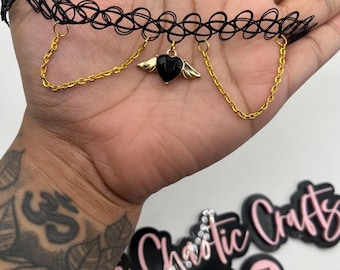 May include: Black choker necklace with a gold chain and a black heart pendant with gold wings. The choker is held in a hand. The image also shows a logo with the text "e Chaotic Crafts".
