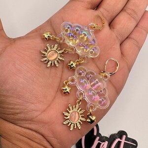 May include: A pair of dangle earrings featuring iridescent, translucent, puffy cloud-shaped charms. Each cloud is accented with a gold-toned sun charm containing a peach-colored center and small gold-toned star charms hanging from the bottom.