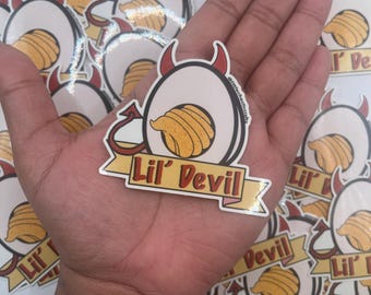 Pode incluir: Um monte de adesivos com um desenho de diabo de desenho animado com um redemoinho amarelo e laranja. Os adesivos têm o texto "Lil' Devil".