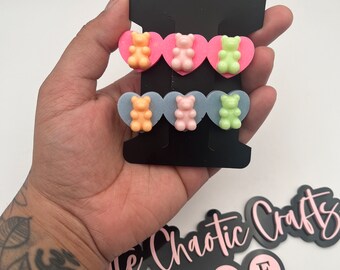 Puede incluir: Un conjunto de seis pendientes de osito de gominola en rosa, naranja y verde. Los pendientes se muestran en una tarjeta negra con un recorte en forma de corazón. La tarjeta también presenta el texto "Little Chaotic Crafts" e iconos de redes sociales.
