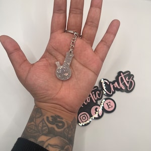 May include: A clear glass bong keychain with silver glitter. The keychain is attached to a silver chain.