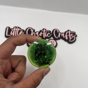 May include: A round, green glittery button with the phrase "This is some Boo Sheet" in black script. The button is held in a hand, with the Little Chaotic Crafts logo in the background.
