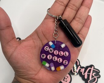 May include: Purple round keyring with the words "CHILL PILLS" spelled out in white letters on a glittery background. The keyring is decorated with colourful pill-shaped charms.