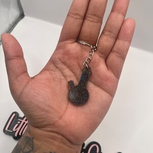 May include: A black and silver glitter bong keychain. The keychain is in the shape of a bong with a round base and a long neck. The keychain is hanging from a silver chain.