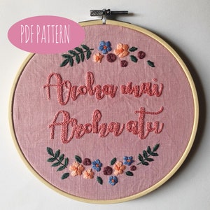 Op de afbeelding: Een roze stoffen borduurring met de woorden "Aroha mai Aroha atu" geborduurd in roze draad. De borduursels zijn versierd met bloemen- en bladmotieven in tinten blauw, oranje, groen en bordeaux. De tekst "PDF PATTERN" staat in een roze ovaal.