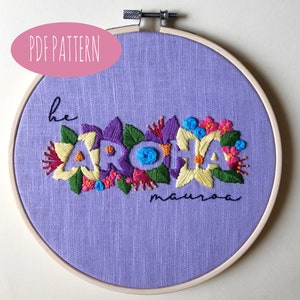 May include: A hand-stitched embroidery hoop featuring the word "ARCHA" in colorful floral designs. The embroidery is on a lavender fabric, with the text "PDF PATTERN" in a pink oval at the top. The word "he" and "mauroa" are also embroidered.