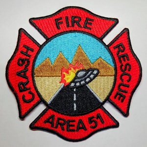May include: Embroidered patch featuring a red Maltese cross design with the words "FIRE", "CRASH", "RESCUE", and "AREA 51". The center depicts a road, mountains, a UFO, and flames, with blue and gold accents.