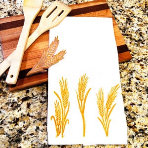 Può includere: Un canovaccio da cucina bianco con un disegno di spighe di grano in inchiostro giallo-arancio. Due utensili da cucina in legno e un tagliere in legno sono sullo sfondo. Il tagliere ha un design a strisce marrone scuro e marrone chiaro.