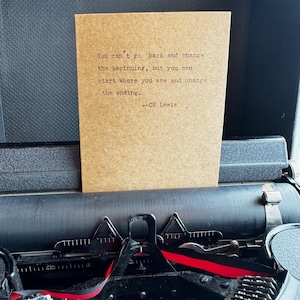 May include: A vintage typewriter with a brown card featuring the quote "You can't go back and change the beginning, but you can start where you are and change the ending." by C.S. Lewis. The typewriter has black keys and red ribbon.
