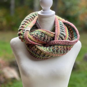 Può includere: Una sciarpa ad anello lavorata all'uncinetto con un motivo di filato variegato nei toni del rosa, verde e marrone. La sciarpa presenta un design testurizzato a coste e include una piccola etichetta rettangolare chiara fissata con un bottone scuro.