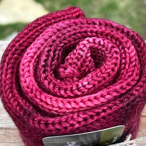 May include: A rolled-up, hand-knitted scarf in shades of deep pink and magenta. The thick, textured knit is tightly coiled, showcasing the intricate stitch work. The scarf appears soft and warm, perfect for cold weather. A tag is visible at the bottom.