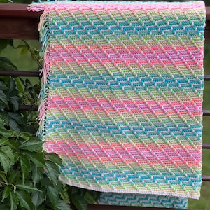 May include: A colorful crocheted blanket with a chevron pattern in shades of pink, green, blue, and white. The blanket has a fringe border and is hanging on a wooden railing.