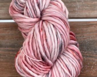 May include: A large ball of chunky yarn in shades of pink, red, and grey. The yarn is thick and textured, ideal for knitting or crochet projects. The yarn is displayed on a wooden surface.