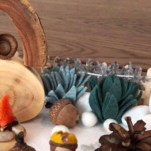May include: A miniature woodland scene with a wooden arch, a felt fire, and two felt figures. The scene is set on a blue felt surface with white felt snow and includes a pine cone, acorns, and white felt bunnies.