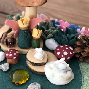 May include: A felt play scene with a wooden mushroom, two small wooden figures with crowns, two white bunnies, a small glass bottle, and various other natural elements.