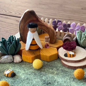 May include: A wooden bee figurine with a white cape and black hair stands on a hexagonal honeycomb. The honeycomb is surrounded by felt flowers, a small glass bottle with glitter, and a wooden archway. The scene is set on a green felt surface.
