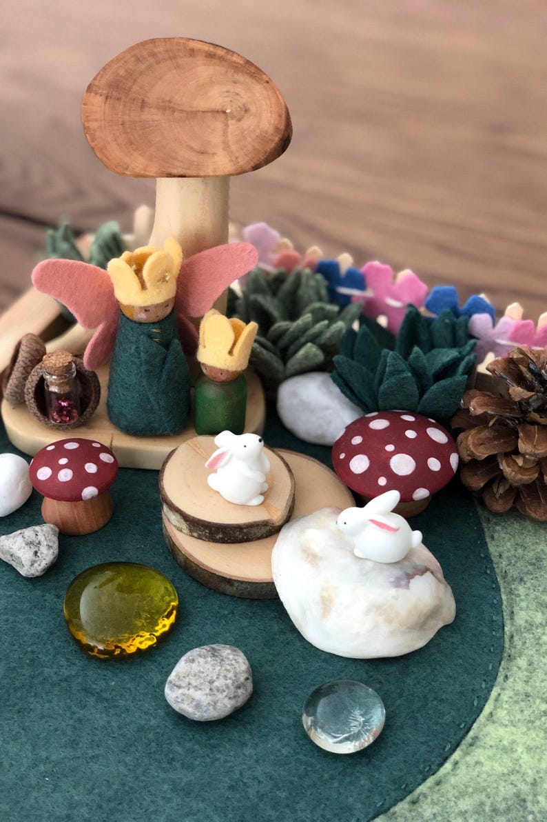 May include: A felt play scene with a wooden mushroom, two small wooden figures with crowns, two white bunnies, a small glass bottle, and various other natural elements.