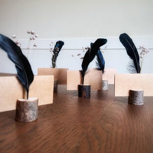 May include: A collection of place card holders with a rustic theme. Each holder features a piece of wood with a black feather and a blank, rectangular card. Delicate, white floral accents are also present.