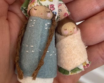 May include: Two handmade wooden peg dolls. One doll is wearing a blue felt dress with white stitching and a floral bonnet. The other doll is wrapped in a pink blanket with a floral trim. Both dolls have painted faces and brown hair.