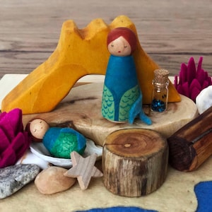 May include: A wooden play set with a yellow wooden arch, two wooden mermaid dolls, a small glass bottle with glitter, a wooden log, a wooden tree stump, and pink felt flowers.