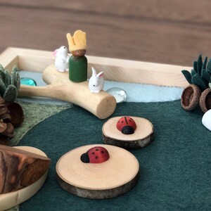 May include: A wooden play set with a green felt base, a wooden tree branch with two white bunny figurines and a green and yellow figurine, two wooden circles with red ladybug figurines, a brown pine cone, a green felt succulent, a white rock, and a red and white mushroom figurine.