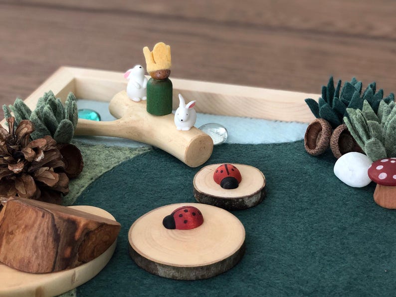 May include: A wooden play set with a green felt base, a wooden tree branch with two white bunny figurines and a green and yellow figurine, two wooden circles with red ladybug figurines, a brown pine cone, a green felt succulent, a white rock, and a red and white mushroom figurine.