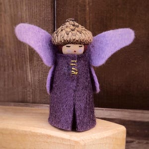 May include: A handmade fairy figure with a felted purple coat and wings. The fairy has an acorn cap head and small gold details on the coat. The figurine is displayed on a wooden block against a wood-panelled backdrop.