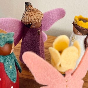 May include: A collection of handmade felt fairy dolls. The fairies have various outfits and accessories, including pink wings, flower crowns, and acorn caps. The dolls are in shades of pink, purple, green, yellow, and white. The dolls are on a wooden surface.